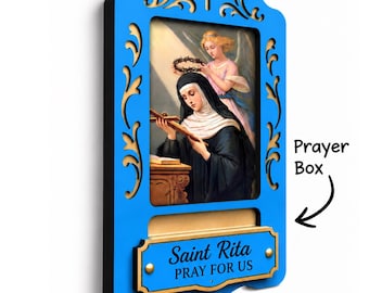 May include: A bright blue wooden wall plaque featuring a central religious image of Saint Rita with an angel, topped with a cross cutout. Below the image is a slot labelled Prayer Box and a plaque with the text Saint Rita PRAY FOR US.