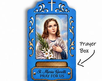 May include: A blue wooden wall plaque featuring a central image of Saint Maria Goretti holding lilies. The frame has a cutout cross at the top and a small prayer box at the bottom with a plaque reading St. Maria Goretti PRAY FOR US.