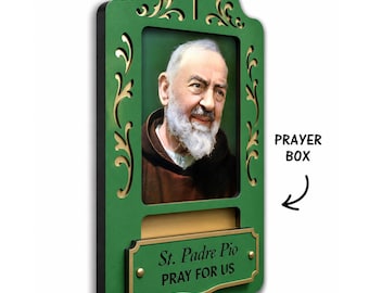 May include: A green wall-mounted prayer box featuring a portrait of St. Padre Pio. The frame has gold-coloured scrollwork and a cross at the top. A gold plaque at the bottom reads St. Padre Pio PRAY FOR US. The text PRAYER BOX is visible.