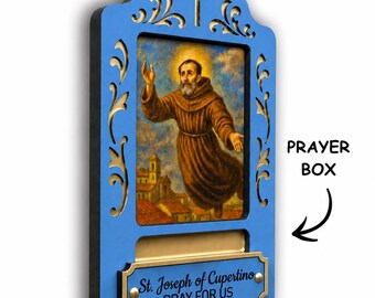 May include: A blue wooden prayer box featuring a central religious painting of St. Joseph of Cupertino. The frame has gold-toned cutouts, a cross at the top, and a plaque reading St. Joseph of Cupertino PRAY FOR US with an arrow pointing to it.