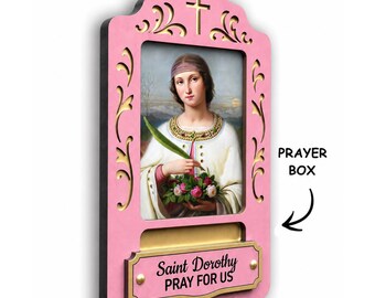 May include: A pink wooden wall-mounted prayer box featuring a central religious portrait of Saint Dorothy holding flowers and a palm leaf. The frame has decorative cutouts, a cross at the top, and a plaque with the text Saint Dorothy PRAY FOR US.