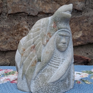 May include: A carved stone sculpture depicting a Native American figure with a wolf headdress. The sculpture is primarily gray with subtle red and white accents. Star-shaped designs are carved into the figure's clothing and headdress.