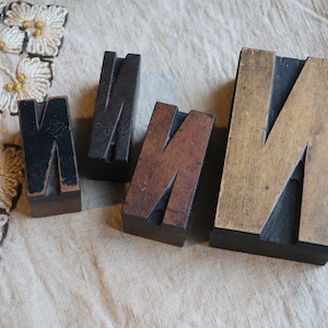 May include: Four wooden letter blocks spelling out the word "MINN" in all caps. The blocks are made of different types of wood and have a worn, vintage look.