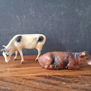 May include: Two small animal figurines. One is a standing white cow with black spots and small horns. The other is a brown cow in a lying position with small horns. Both figures are painted with detailed features and are positioned side by side.
