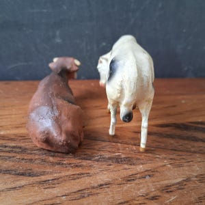 May include: Two small animal figurines, one solid brown cow in a lying position and one white cow with black spots standing upright. Both figures are viewed from the rear, showing their tails and body shapes in a detailed, realistic style.