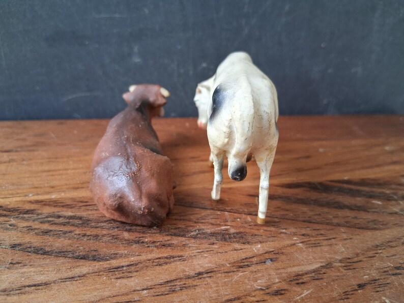 May include: Two small animal figurines, one solid brown cow in a lying position and one white cow with black spots standing upright. Both figures are viewed from the rear, showing their tails and body shapes in a detailed, realistic style.