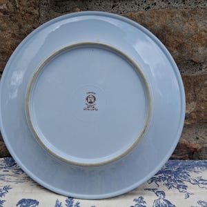 Pu&ograve; includere: Un piatto in ceramica azzurra, visto dal basso, con un design circolare e un bordo marrone. Il piatto ha un timbro al centro. Il piatto &egrave; su un tessuto a fantasia blu e bianco.
