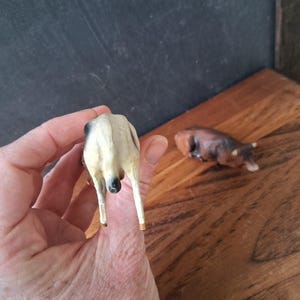 May include: A small, standing figurine of a white cow with black markings, viewed from the rear, alongside a separate, smaller, brown cow figurine lying down in a resting position. Both figures are crafted with detailed textures.