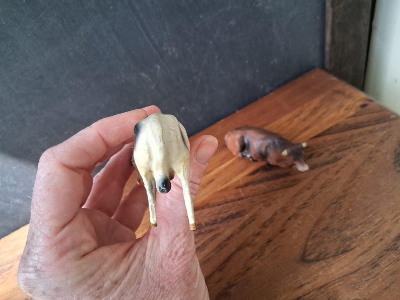 May include: A small, standing figurine of a white cow with black markings, viewed from the rear, alongside a separate, smaller, brown cow figurine lying down in a resting position. Both figures are crafted with detailed textures.
