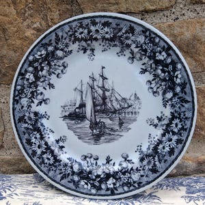 Pu&ograve; includere: Un piatto decorativo bianco e blu con una scena nautica al centro. Il piatto presenta un'illustrazione dettagliata di una nave alta che naviga sull'acqua, circondata da un bordo floreale. Il piatto &egrave; probabilmente in ceramica.