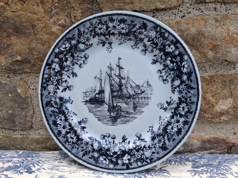 Pu&ograve; includere: Un piatto decorativo bianco e blu con una scena nautica al centro. Il piatto presenta un'illustrazione dettagliata di una nave alta che naviga sull'acqua, circondata da un bordo floreale. Il piatto &egrave; probabilmente in ceramica.