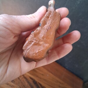 May include: A small, hand-sculpted clay figurine with a glossy, reddish-brown finish. The object has an irregular, organic shape with a smooth, reflective surface texture and a small, rounded, light-colored protrusion at the top end.