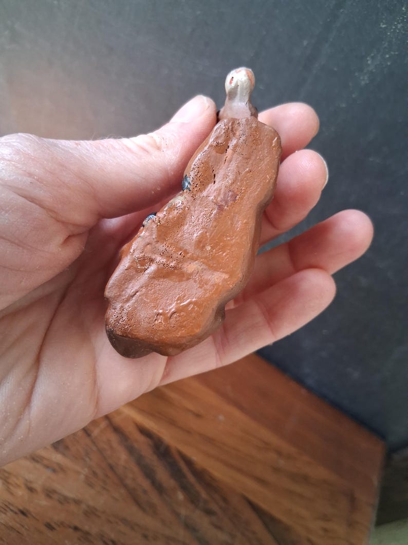 May include: A small, hand-sculpted clay figurine with a glossy, reddish-brown finish. The object has an irregular, organic shape with a smooth, reflective surface texture and a small, rounded, light-colored protrusion at the top end.