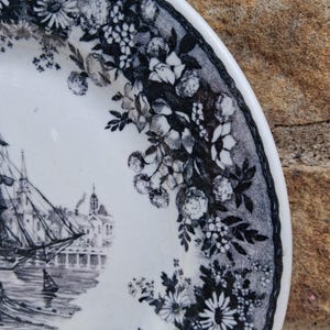 Pu&ograve; includere: Piatto in ceramica bianca con una scena nautica in bianco e nero con una nave alta, edifici e un bordo floreale. Il piatto ha un design dettagliato e un'estetica classica.