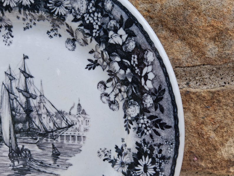 Pu&ograve; includere: Piatto in ceramica bianca con una scena nautica in bianco e nero con una nave alta, edifici e un bordo floreale. Il piatto ha un design dettagliato e un'estetica classica.