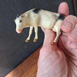 May include: A small, off-white cow figurine featuring black patches on its head and rear. The animal has a pink-toned nose and brown-painted hooves. The figure is sculpted in a standing position with four legs and visible ears, showcasing a smooth, matte finish.