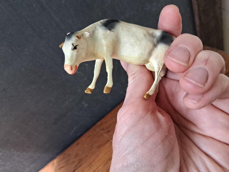 May include: A small, off-white cow figurine featuring black patches on its head and rear. The animal has a pink-toned nose and brown-painted hooves. The figure is sculpted in a standing position with four legs and visible ears, showcasing a smooth, matte finish.