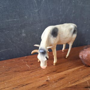 May include: A small, standing figurine of a white cow with black spots on its back and head. The cow has curved horns, a light-colored muzzle, and is positioned in a downward-facing pose. A portion of a brown animal figurine is visible to the right.