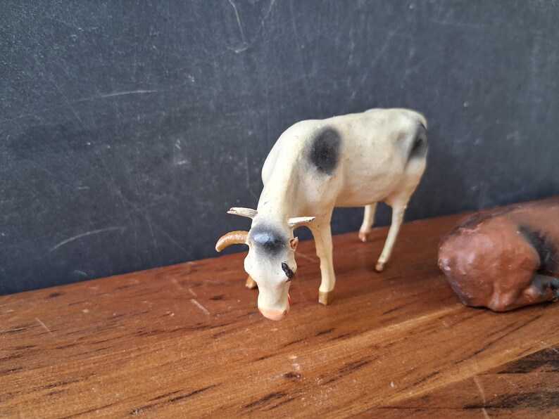 May include: A small, standing figurine of a white cow with black spots on its back and head. The cow has curved horns, a light-colored muzzle, and is positioned in a downward-facing pose. A portion of a brown animal figurine is visible to the right.