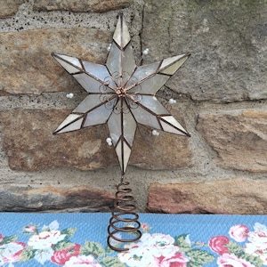 May include: A six-pointed star tree topper featuring iridescent white panels framed in dark copper metal. The center is adorned with intricate copper wire filigree and small white beads are attached to the points of the star. A copper wire coil base.