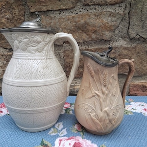 May include: Two ceramic pitchers with metal lids. The larger pitcher is cream-colored with a textured basket-weave pattern and embossed fruit motifs. The smaller pitcher is tan with an embossed wheat and floral design and a curved handle.