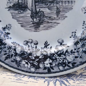 Pu&ograve; includere: Un piatto in ceramica bianca con una scena nautica blu e bianca al centro. Il bordo del piatto presenta un motivo floreale con margherite e altri fiori. Il piatto &egrave; probabilmente vintage o antico.