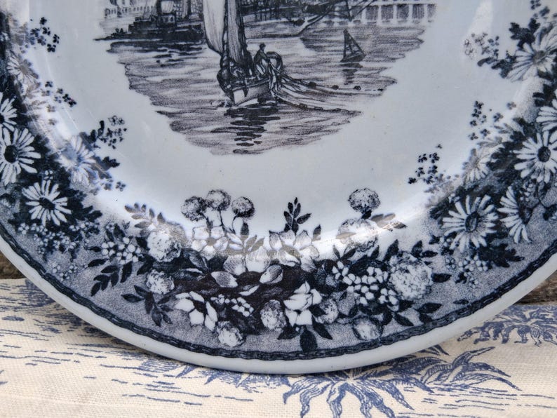 Pu&ograve; includere: Un piatto in ceramica bianca con una scena nautica blu e bianca al centro. Il bordo del piatto presenta un motivo floreale con margherite e altri fiori. Il piatto &egrave; probabilmente vintage o antico.