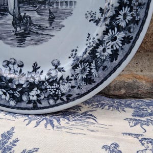 Pu&ograve; includere: Un piatto in ceramica bianca con una scena nautica blu e bianca e un bordo floreale. Il piatto presenta un'illustrazione dettagliata di una barca a vela sull'acqua. Il bordo &egrave; decorato con fiori e foglie blu. Il piatto &egrave; appoggiato su un tessuto a fantasia blu e bianco.