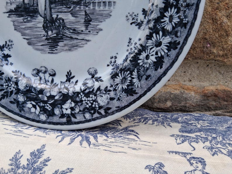 Pu&ograve; includere: Un piatto in ceramica bianca con una scena nautica blu e bianca e un bordo floreale. Il piatto presenta un'illustrazione dettagliata di una barca a vela sull'acqua. Il bordo &egrave; decorato con fiori e foglie blu. Il piatto &egrave; appoggiato su un tessuto a fantasia blu e bianco.