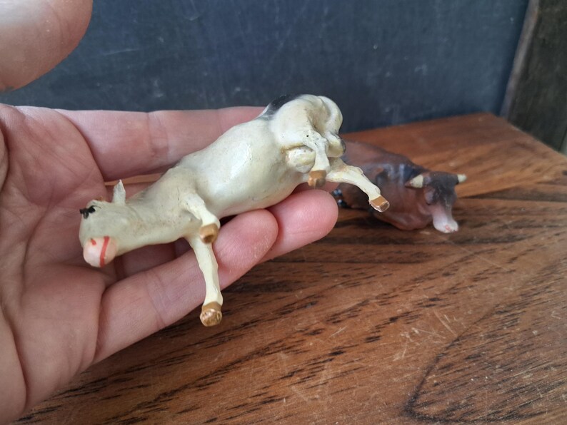 May include: Two small animal figurines, one cream-colored cow with black markings and one brown cow, both depicted in a lying position. The cream cow is held in the palm of a hand, while the brown cow is positioned on a wooden surface nearby.