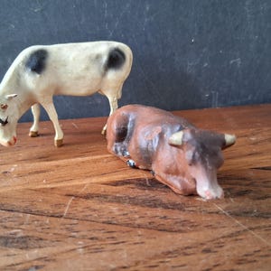 May include: Two small animal figurines consisting of a standing white cow with black spots and a brown cow in a lying position. Both figures feature painted details including horns, ears, and facial features, crafted as decorative miniature livestock.