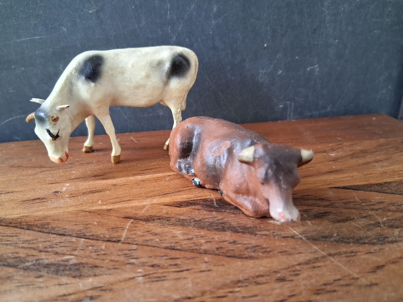May include: Two small animal figurines consisting of a standing white cow with black spots and a brown cow in a lying position. Both figures feature painted details including horns, ears, and facial features, crafted as decorative miniature livestock.