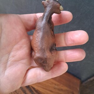 May include: A small, dark brown, textured figurine of a four-legged animal with a slender neck and two light-colored, pointed horns on its head. The object has a matte finish and a curved, organic shape, appearing to be crafted from clay or stone.