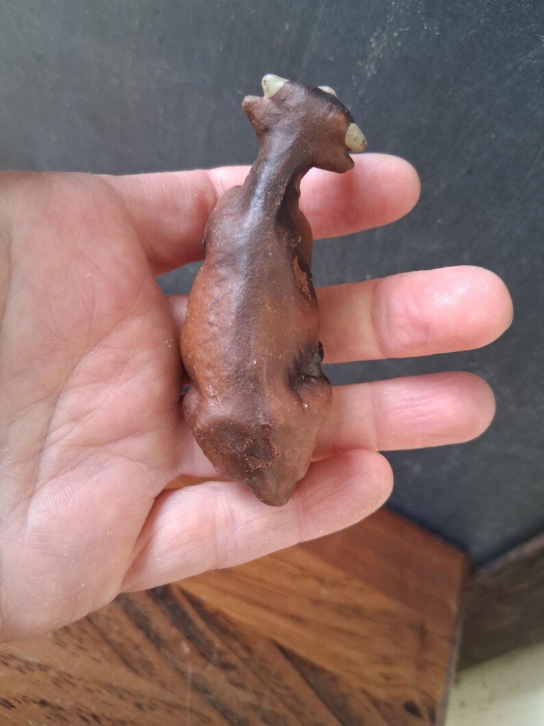 May include: A small, dark brown, textured figurine of a four-legged animal with a slender neck and two light-colored, pointed horns on its head. The object has a matte finish and a curved, organic shape, appearing to be crafted from clay or stone.