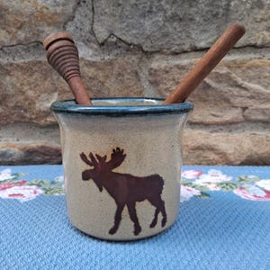 May include: A beige ceramic honey pot featuring a dark brown moose silhouette on the front. The pot has a dark blue rim and contains a wooden honey dipper and a wooden stirring stick, both extending from the top opening of the container.