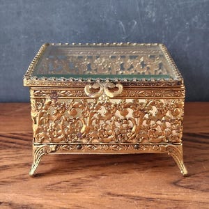 May include: A rectangular jewelry box featuring a gold-toned metal frame with intricate floral filigree patterns, a clear glass lid, and four small decorative legs. The box has a hinged top with a central metal clasp and a green interior lining.