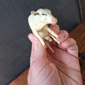May include: A small, off-white cow figurine with a black patch on its head and gold-painted accents on the horns and hooves. The miniature animal is depicted in a seated position with a pink-toned snout and visible facial features.