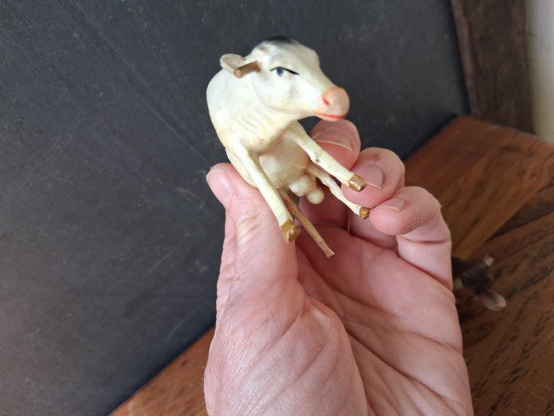 May include: A small, off-white cow figurine with a black patch on its head and gold-painted accents on the horns and hooves. The miniature animal is depicted in a seated position with a pink-toned snout and visible facial features.