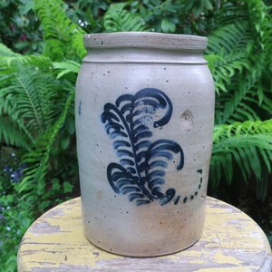 Pode incluir: Um pote de barro vintage com um design de samambaia azul e branco. O pote está sentado em um banquinho pintado de amarelo.