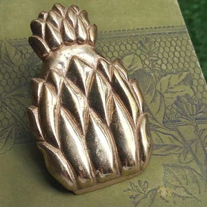 May include: A polished brass object shaped like a pineapple with a textured, segmented surface. The metallic piece features a rounded top section and a wider, tapered base with distinct, raised diamond-shaped patterns across its entire body.