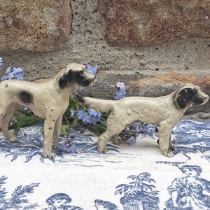 May include: Two antique dog figurines. One is tan with a black spot, the other is tan with black markings. They are posed on a blue and white patterned fabric, with blue flowers in the background. The dogs are made of a light-colored material.