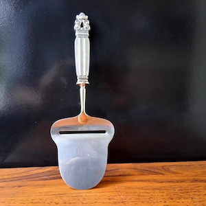 May include: A silver cheese slicer with a decorative handle and a thin blade. The handle has a detailed design at the top and is connected to the blade by a thin, gold-colored stem. The blade has a curved shape with a slot.