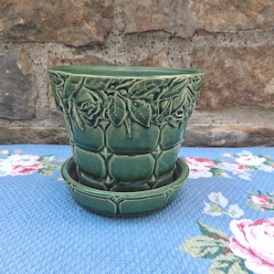 May include: A glossy forest green ceramic plant pot with a matching saucer. The exterior features a raised relief pattern of rose blossoms and leaves around the rim, transitioning into a structured grid of square panels along the lower body.