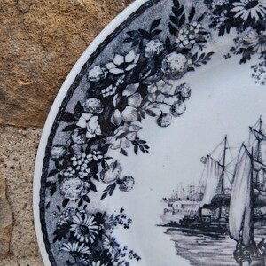 Pu&ograve; includere: Piatto in ceramica bianca con bordo floreale in bianco e nero e scena nautica. Il piatto presenta un'illustrazione dettagliata di velieri e una nave a vapore sull'acqua. Il design floreale include margherite e altri fiori.
