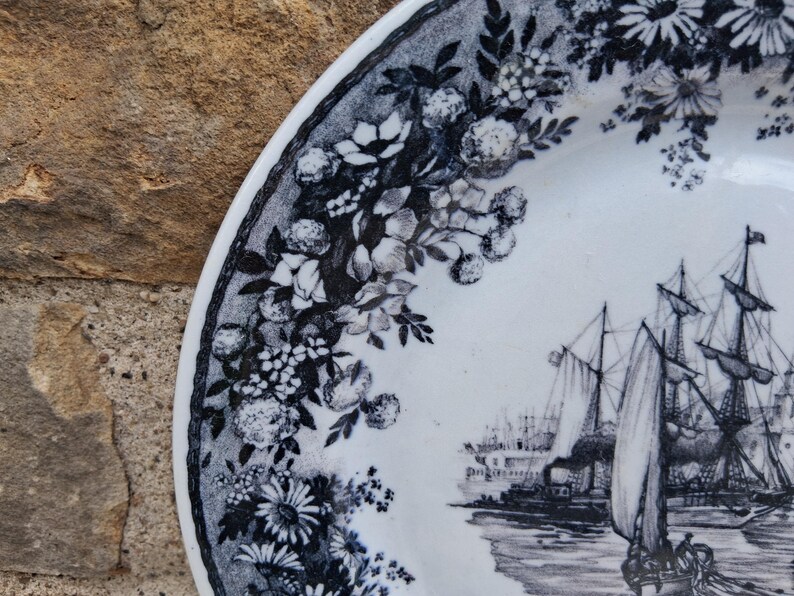 Pu&ograve; includere: Piatto in ceramica bianca con bordo floreale in bianco e nero e scena nautica. Il piatto presenta un'illustrazione dettagliata di velieri e una nave a vapore sull'acqua. Il design floreale include margherite e altri fiori.