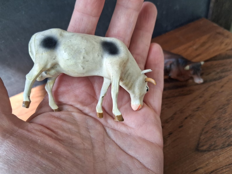 May include: A small, off-white cow figurine with two prominent black spots on its back. The miniature animal features light brown hooves, small horns, and a pinkish snout, depicted in a grazing position with its head lowered toward the ground.