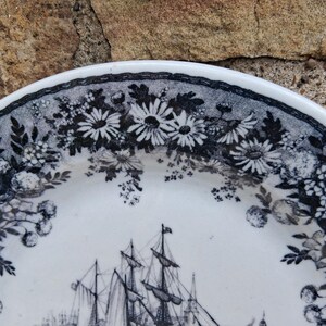 Pu&ograve; includere: Piatto in ceramica bianca con bordo floreale bianco e nero e un'illustrazione dettagliata di una nave a vela. Il design del piatto presenta margherite e altri fiori.