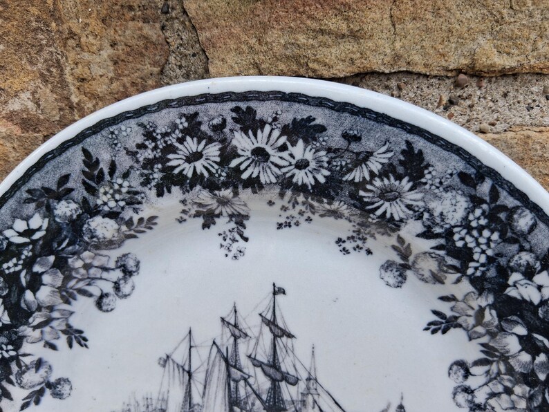 Pu&ograve; includere: Piatto in ceramica bianca con bordo floreale bianco e nero e un'illustrazione dettagliata di una nave a vela. Il design del piatto presenta margherite e altri fiori.