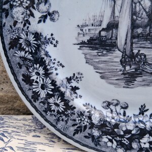 Pu&ograve; includere: Un piatto in ceramica bianca con un bordo floreale blu e bianco e una scena nautica. Il piatto presenta un'illustrazione dettagliata di barche a vela e di un porto. Il bordo floreale comprende margherite e altri fiori.