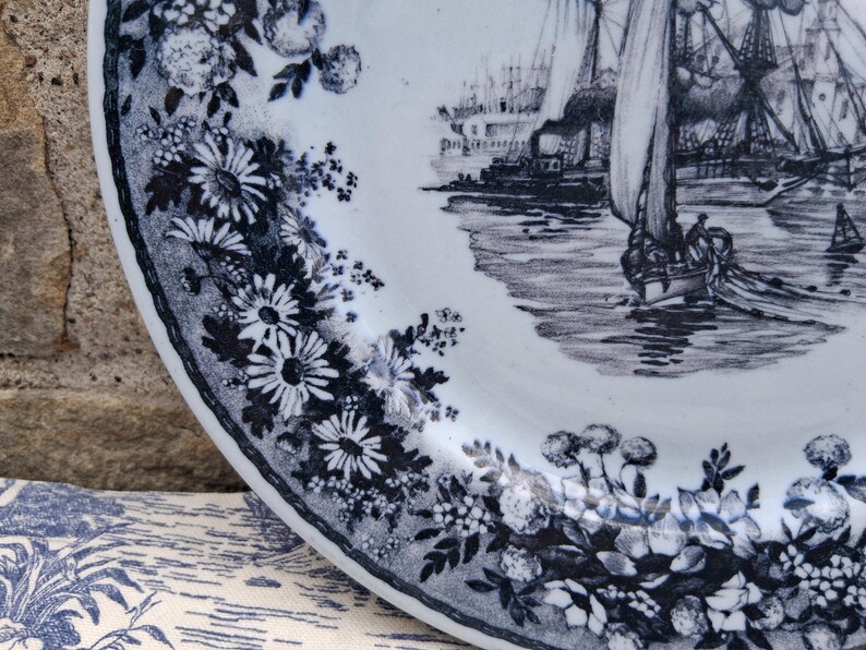 Pu&ograve; includere: Un piatto in ceramica bianca con un bordo floreale blu e bianco e una scena nautica. Il piatto presenta un'illustrazione dettagliata di barche a vela e di un porto. Il bordo floreale comprende margherite e altri fiori.